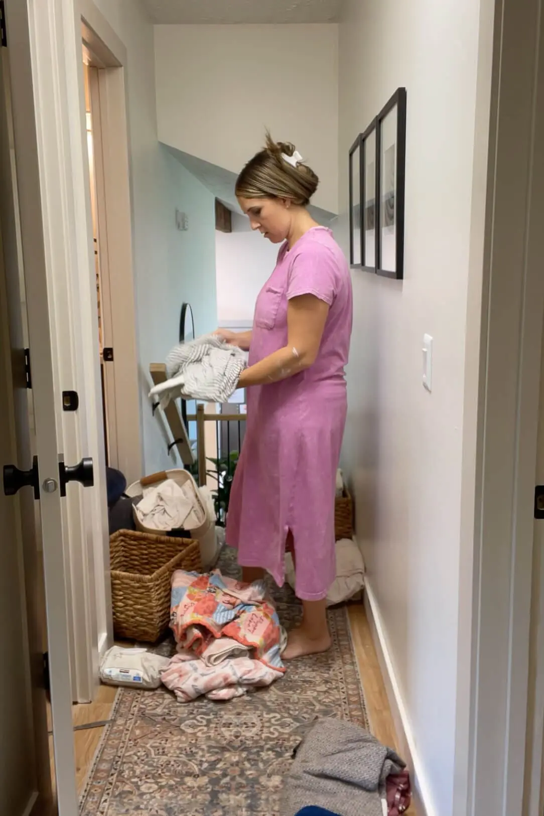 Woman in a pink dress sorting through a pile of sheets, blankets, and linens in a hallway before reorganizing a linen closet.